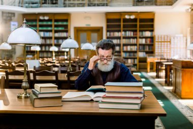 Portrait of elegant senior man, university school teacher, wruter, with beard and eyeglasses, reading books in vintage ancient library on bookshelf background. Books and vintage desk lamp on the table