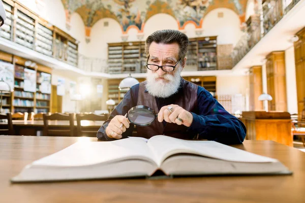 Γέρο επιστήμονα, βιβλιοθηκάριο, διάβαζε ένα βιβλίο σε μια βιβλιοθήκη, κοιτάζοντας μέσα από το μεγεθυντικό φακό. Vintage ράφια βιβλιοθήκη και βιβλία στο παρασκήνιο — Φωτογραφία Αρχείου