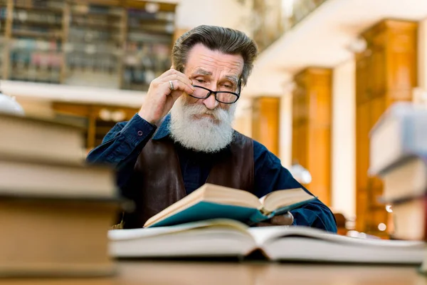 Elderly old man professor, writer, reading a book in old vintage library. An old intelligent man wearing eyeglasses and stylish clothes reading a book in city library