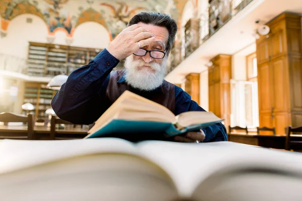 Ανώτερος συμπυκνωμένος άνδρας με λευκή γενειάδα, αγγίζει τα γυαλιά του, κάθεται στο τραπέζι και σκέφτεται το βιβλίο που διαβάζει στη βιβλιοθήκη του πανεπιστημίου — Φωτογραφία Αρχείου