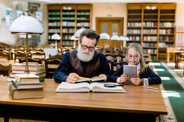 Cute little girl uses the application on the tablet, and an elderly man reads a book in the library. Old and new technologies, paper book and ebook, grandfather and granddaughter — Φωτογραφία Αρχείου