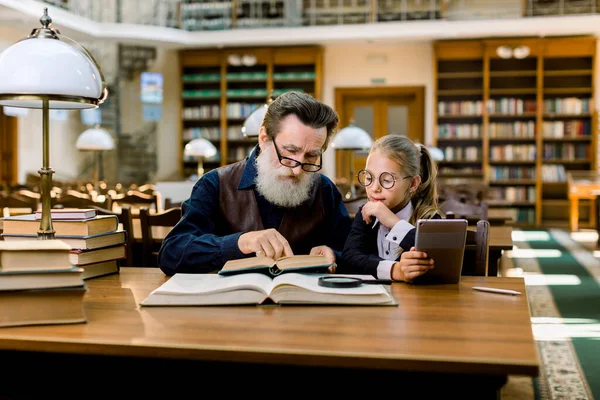 Profesor principal profesor enseñando a una niña pequeña, mostrándole un libro, mientras está sentado en la biblioteca con un fondo de librerías vintage. Chica sostiene la tableta y mirando el libro —  Fotos de Stock