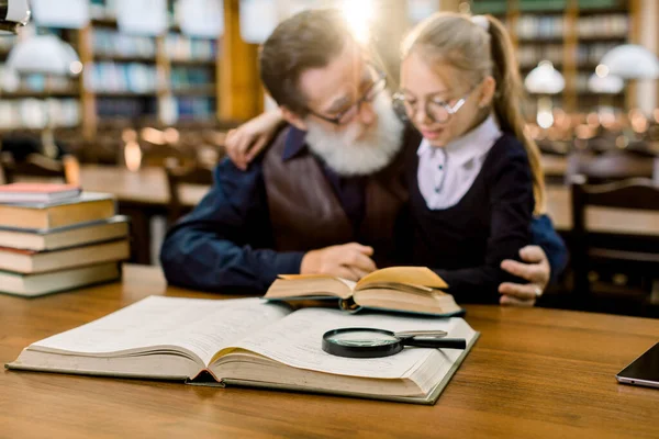 Abuelo y nieta leyendo un libro en la antigua biblioteca de la ciudad. Lectura familiar, ocio, concepto educativo. Enfócate en los libros y la lupa en la mesa — Foto de Stock