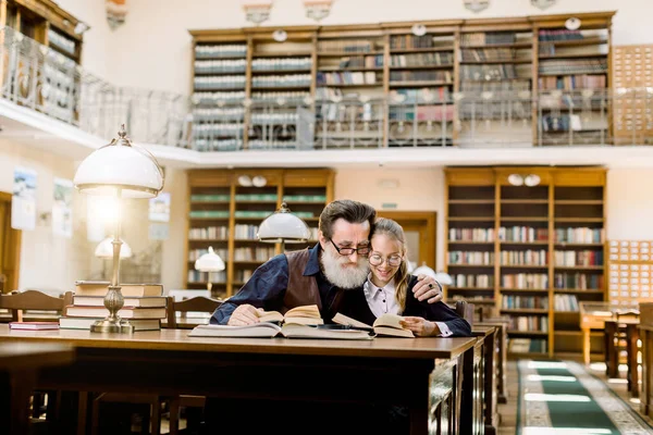 Ένα μικρό κορίτσι και ο μεγαλύτερος γενειοφόρος παππούς της διαβάζουν βιβλία, κάθονται στο τραπέζι με πολλά βιβλία και vintage λάμπα γραφείου στην παλιά αρχαία βιβλιοθήκη, στο παρασκήνιο των βιβλιοθηκών — Φωτογραφία Αρχείου