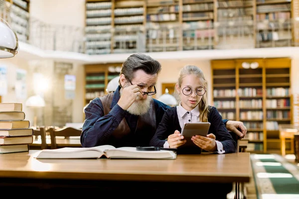 Starší muž učitel se svou malou studentskou žákyní používá digitální tablet, sedí spolu u stolu ve staré starověké knihovně. — Stock fotografie
