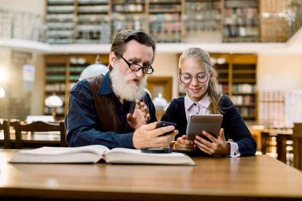 Abuelo y nieta, maestro y estudiante, sentados juntos en la mesa usando tableta digital, smartphone y libros. Estudio, lectura familiar en la biblioteca. Concepto educativo — Foto de Stock