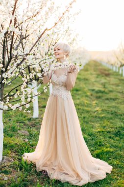 Çiçekli bir bahar bahçesinde çiçek açan meyve ağacının yanında poz veren uzun ve zarif elbiseli güzel, genç bir gelin.