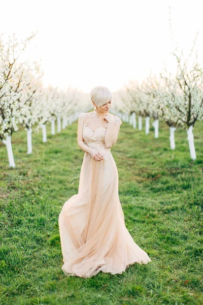 Portrait de jeune jolie femme blonde en robe élégante de mariage dans le jardin fleuri au printemps. Des arbres fruitiers fleurissent, des lignes d'arbres sur le fond — Photo