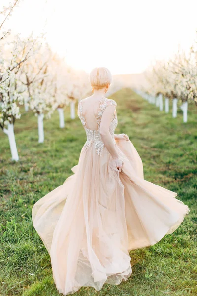 Jeune belle romantique blonde femme caucasienne en longue robe élégante lumière marche et courir dans le jardin en fleurs, vue de dos portrait pleine longueur — Photo