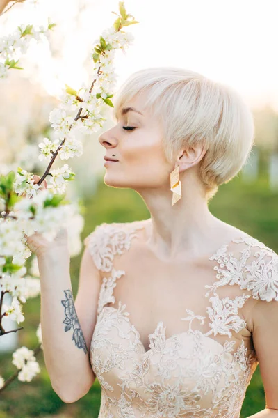 Portrait de mode de belle femme avec des cheveux blonds courts dans une robe de mariée luxueuse posant dans le jardin de printemps en fleurs, toucher branche d'arbre avec des fleurs en fleurs — Photo