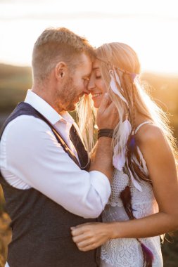 beautiful couple dressed in boho style embracing each other in the sunny summer field. Handsome man in casual wear and woman in white dress and feathers in hair walking in the field at sunset