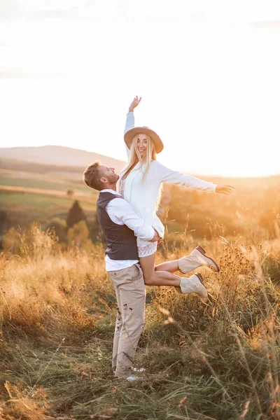 Casal jovem vestindo roupas estilo hipster boho indie, no amor andando no campo, o homem mantém a mulher a mãos, a mulher está feliz e sorrindo. Campo de pôr-do-sol ensolarado no fundo, dia quente de verão — Fotografia de Stock