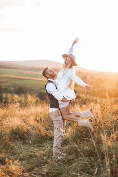 Genç çift hipster boho tarzı indie kıyafetleri giyiyor, kırsalda aşık yürüyor, erkek kadını elinde tutuyor, kadın mutlu ve gülümsüyor. Güneşli günbatımı tarlası arka planda, sıcak yaz günü