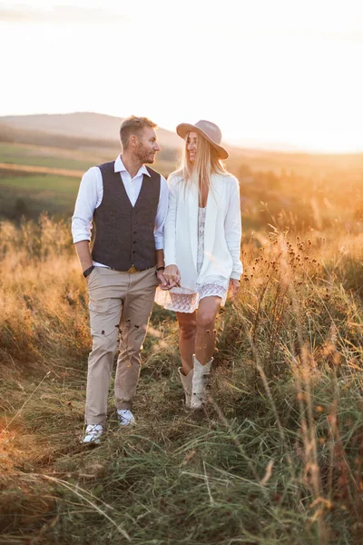 Bonito homem elegante em terno rústico e mulher boho bonita em vestido, jaqueta, chapéu e botas de cowboy, andando no campo, de mãos dadas, olhando um para o outro e sorrindo. Noite de verão, pôr do sol — Fotografia de Stock