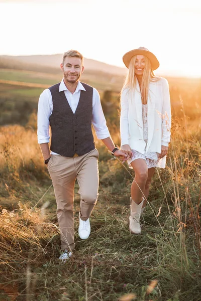 Verão, amor, romance e conceito de pessoas - feliz sorrindo jovem casal estilo cowboy hippie de mãos dadas e andando no campo de verão, ao ar livre. Mulher de vestido e botas de cowboy, homem de terno casual — Fotografia de Stock