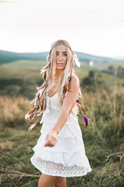 Mulher boho loira atraente feliz em vestido branco e acessórios de penas no cabelo no campo de verão ao ar livre, pôr do sol. Boho moda, estilo hippie — Fotografia de Stock