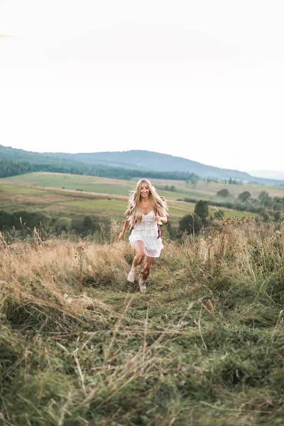 Feliz mulher sorridente com cabelo loiro longo vestindo vestido hippie boho branco se divertindo enquanto corre em campo. Penas no cabelo, estilo boho — Fotografia de Stock