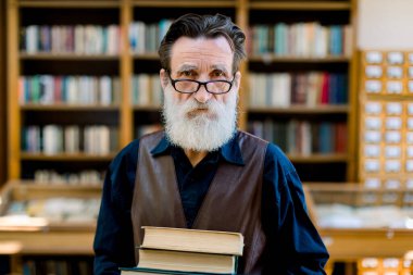 Close up portrait of handsome bearded senior academic professor or library worker, smiling and holding old books, while standing over the background of vintage library bookcases. Knowledge concept clipart