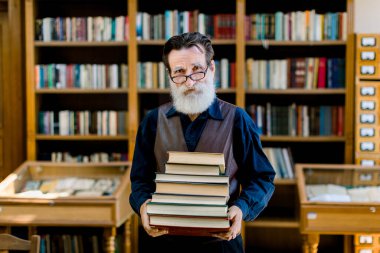Portrait of positive smart old bearded man in dark shirt and leather vest, library worker, teacher, working in library, holding stack of books while standing over book shelves background clipart