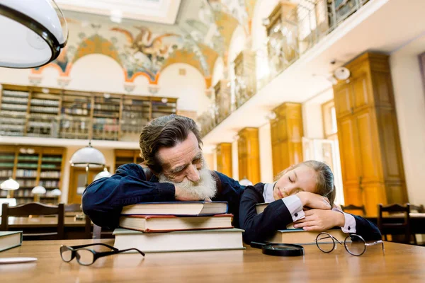 Unavený starší vousatý muž učitel profesor a jeho student nebo vnučka spí v knihovně ležící na stole se spoustou knih ve staré archivní knihovně — Stock fotografie