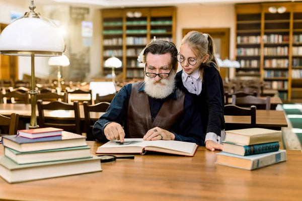 Anciano abuelo hombre y su nieta leer libro emocionante juntos en la antigua biblioteca de la vendimia, en el fondo de los estantes de libros. Lámpara de escritorio vintage y libros sobre la mesa . —  Fotos de Stock