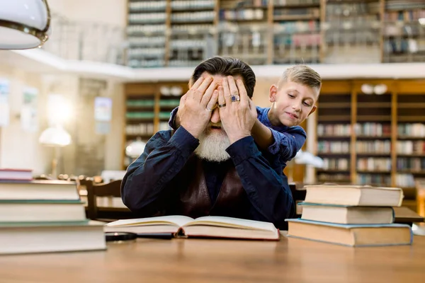 Niespodzianka. Szczęśliwego dnia książki, rodzinnego pomysłu. Mały wnuczek zamyka rękami oczy dziadka, siedzi przy stole i czyta książki w zabytkowej bibliotece. Zgadnij, kto tam jest.. — Zdjęcie stockowe