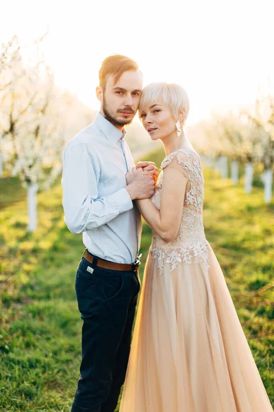 Jovem bela mulher casal e homem vestindo roupas elegantes de luxo, de mãos dadas e abraços, posando ao ar livre sobre florescendo árvores fundo, dia de primavera ensolarado e pôr do sol — Fotografia de Stock