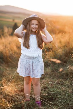 Beyaz elbiseli ve kahverengi şapkalı şirin küçük kız gün batımında yaz tarlasında duruyor. Kırsal hayat, tarlada yürü.