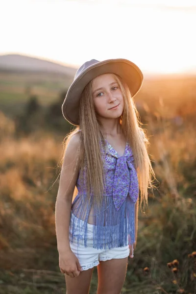 Petite fille mignonne aux longs cheveux blonds, coiffée d'un chapeau marron, posant dans le champ de fleurs et d'herbes sauvages avec coucher de soleil — Photo