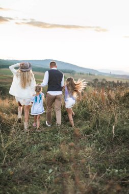 Moda kıyafetleri içinde neşeli bir ailenin arka plan fotoğrafı, ebeveynler ve çocuklar, dağlarda birlikte koşup eğleniyorlar. Aile kırsalda, açık havada.