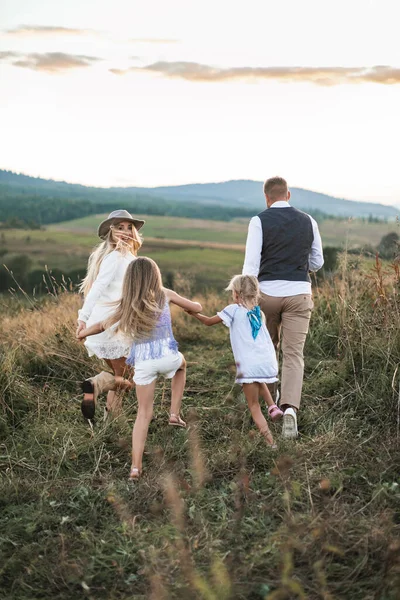Hora de verão no campo. Visão traseira da família com duas filhas pequenas correndo e se divertindo no campo selvagem de verão ao pôr do sol . — Fotografia de Stock