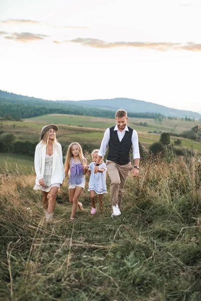 Gündelik giysiler içinde mutlu bir aile, anne, baba ve iki küçük kız, el ele yürüyorlar, dağlardaki vahşi tarlalarda gün batımını izliyorlar. — Stok fotoğraf