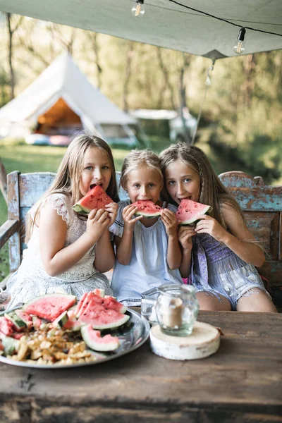 Drei süße, glücklich lächelnde Mädchen, Schwestern, Teufelinnen, die am Tisch auf einer alten Holzbank sitzen und draußen im Zelt Wassermelone essen, im Hintergrund stylische Boho-Perücken. Sommerferien auf dem Land — Stockfoto