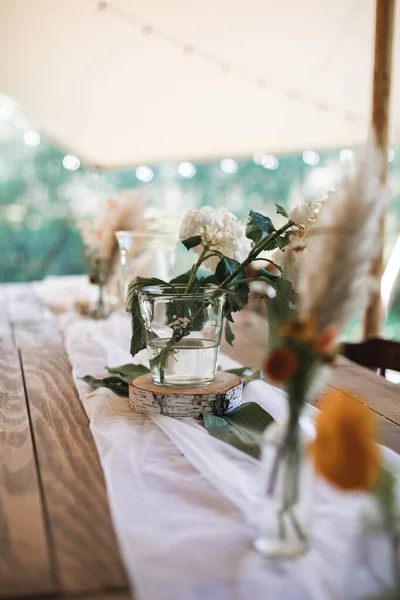 Rustykalne dekoracje stołowe na zewnątrz. Bukiety kwiatów, suche dzikie kwiaty w szklanych słoikach na drewnianej wannie leżącej na świątecznym drewnianym stole, stole weselnym lub obiadowym na zewnątrz — Zdjęcie stockowe
