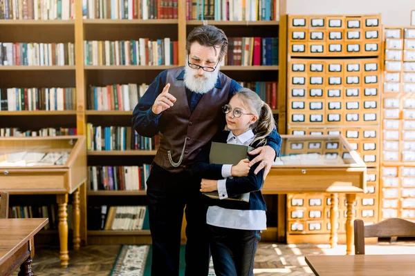 Happy excited teen blond girl with glasses holding a book in hands while listening her intelligent old grandpa, teacher or librarian, showing the library interior and talking about interesting books