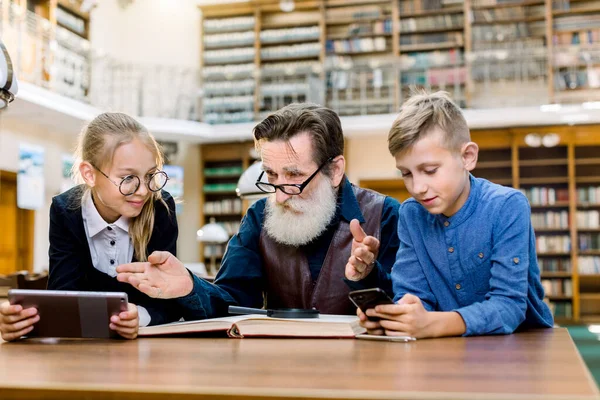 Petits enfants souriants d'âge scolaire, garçon et fille, comparant tablette e-reader et smartphone avec leurs grands-pères livre traditionnel. Homme barbu âgé essaie de convaincre les enfants de lire un livre — Photo