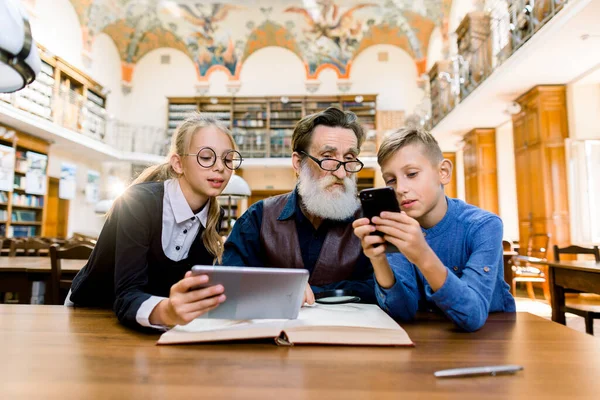 Technologie, ordinateur par rapport au concept traditionnel des livres imprimés. Grand-père et petits-enfants sont assis à la table de la bibliothèque. Enfants apprenant à son grand-père à utiliser une tablette numérique et un smartphone . — Photo