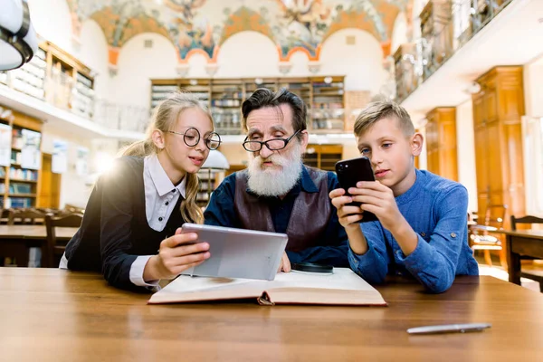 Pequena neta e neto ensinar o avô idoso sênior para navegar na internet usando tecnologias modernas, tablet digital e smartphone. Avô feliz com netinhos sentados na biblioteca — Fotografia de Stock