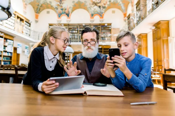 Os netos felizes estudam juntos com seu avô, fazendo trabalhos de casa na biblioteca, lendo livros e usando tablet e smartphone para obter informações. Conceito de aprendizagem . — Fotografia de Stock