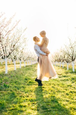 Romantik genç adam kadınını kollarında taşırken sevgi dolu bir tebessümle gözlerinin içine bakarken güzel çiçek açan bahçedeki yeşil çimlerin üzerinde dikilirken.