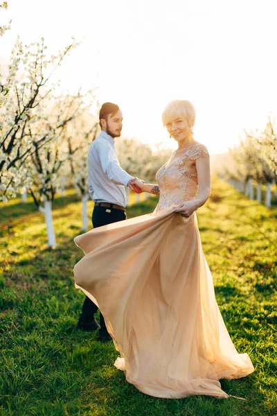 Frühlingsporträt eines schönen glücklichen Paares, das im Frühling blühenden Garten tanzt und Zeit verbringt. Lächelnde Frau mit kurzen blonden Haaren in exquisitem Kleid blickt in die Kamera — Stockfoto