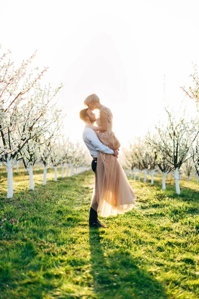 Молодая пара в цветущем весеннем саду сливы. Счастливый красивый мужчина, несущий свою красавицу жену, целующий его в лоб, стоящий между красивыми цветущими деревьями на фоне заката — стоковое фото