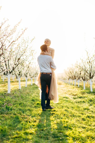 Tiro de encantadora mulher loira muito jovem em vestido de noiva de luxo sendo levado por seu homem bonito no jardim florescendo primavera. Casal se divertindo em sua caminhada de casamento — Fotografia de Stock