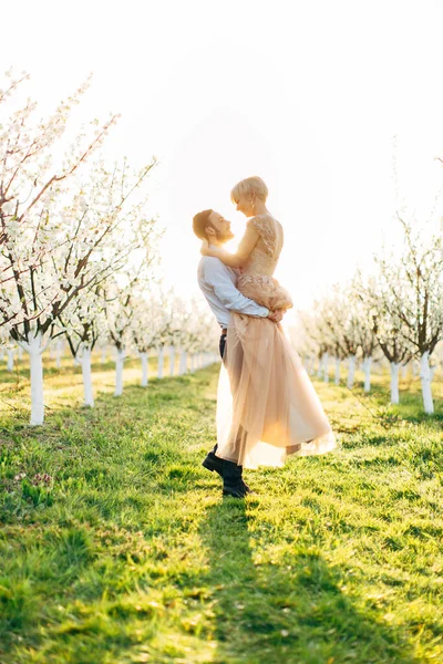 Романтичный молодой человек, несущий свою женщину на руках, с любящей улыбкой глядя ей в глаза, стоя на зеленой траве в красивом цветущем саду — стоковое фото