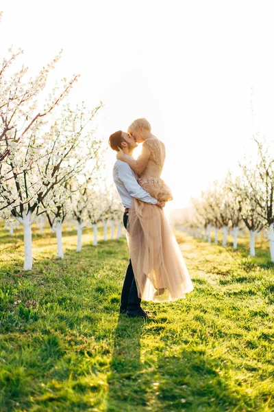 Beau jeune couple sensuel s'embrassant dans un endroit romantique, jardin de pommes fleurissant au printemps. Heureux couple joyeux profiter l'un de l'autre pendant le lever ou le coucher du soleil dans le jardin. Homme tenant femme sur les mains — Photo