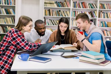 Üniversite kütüphanesindeki masada oturup sınav hazırlıkları sırasında dizüstü bilgisayar kullanan ve kitap okuyan neşeli, çok kültürlü öğrenciler.