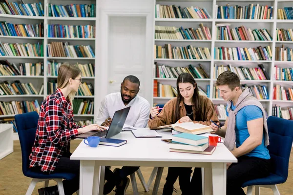 Concepto de educación y aprendizaje. Grupo multiétnico de estudiantes universitarios diversos y concentrados que se sientan a la mesa y se preparen juntos para los exámenes en la biblioteca moderna —  Fotos de Stock