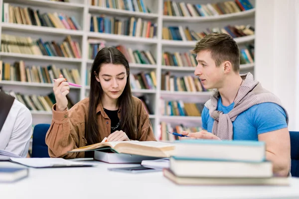 Dois atraentes e sorridentes estudantes confiantes, caucasianos menina e menino, sentados à mesa enquanto se preparam juntos para exames na biblioteca usando diferentes livros, conversando uns com os outros — Fotografia de Stock