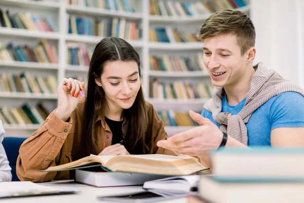 Étudiants, beau garçon caucasien et jolie fille, assis à une table dans une bibliothèque tout en lisant des livres et en apprenant ensemble, en discutant de leur travail à domicile ou de la préparation à l'examen. Éducation, amitié — Photo