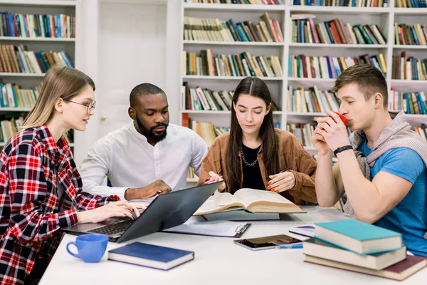 Vista frontal de cuatro estudiantes de raza mixta moderna y atractiva que están sentados en la sala de lectura de la biblioteca y preparándose para los exámenes o el proyecto científico. Libros, portátil, tableta sobre la mesa — Foto de Stock